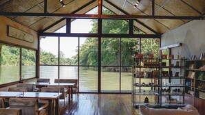 A spacious dining area with wooden tables and chairs, large windows overlooking a river, and shelves with various items. - Long Chuan River Kwai (Thong Pha Phum)