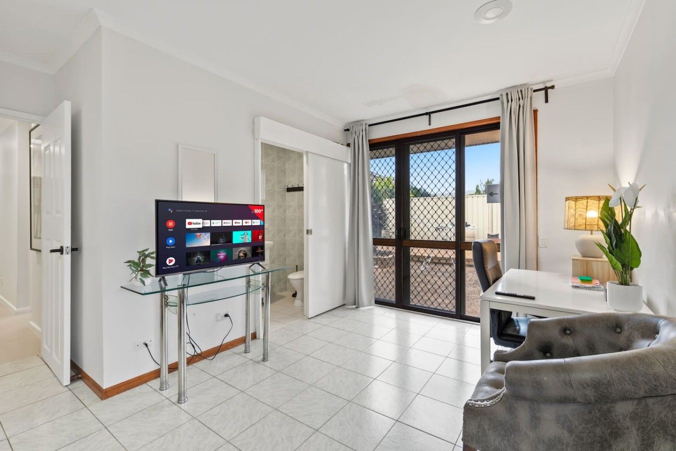 A modern living room with a flat-screen TV on a glass stand, a grey sofa, a dining table with a lamp, and a sliding glass door leading to a balcony.