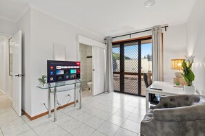 A modern living room with a flat-screen TV on a glass stand, a grey sofa, a dining table with a lamp, and a sliding glass door leading to a balcony. - Affordable 1-Bed Studio Close to Malls & Transit! (Robertson)