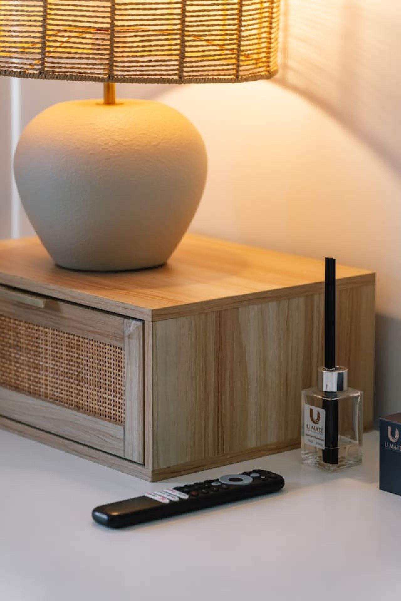 A bedside table with a lamp, a remote control, and a bottle of perfume.