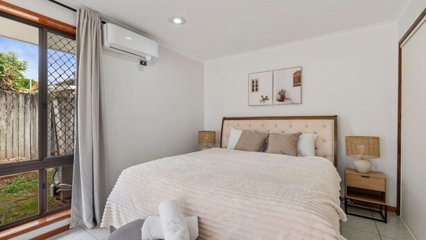 A bedroom with a large bed, a bench, a nightstand, and a window with a view of the outdoors. - Affordable 1-Bed Studio Close to Malls & Transit! (Robertson)