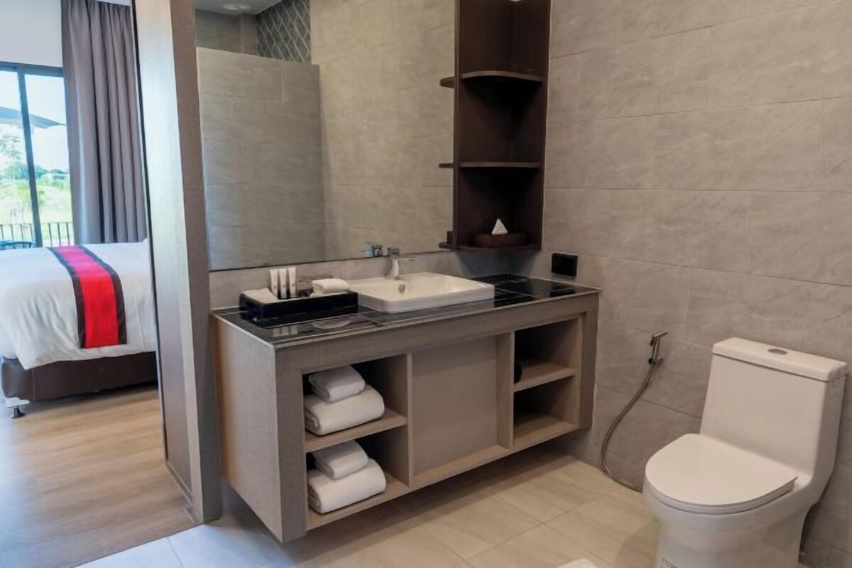 A modern bathroom with a large mirror, a white sink, and a toilet. There is a bed with a red blanket in the adjacent room.