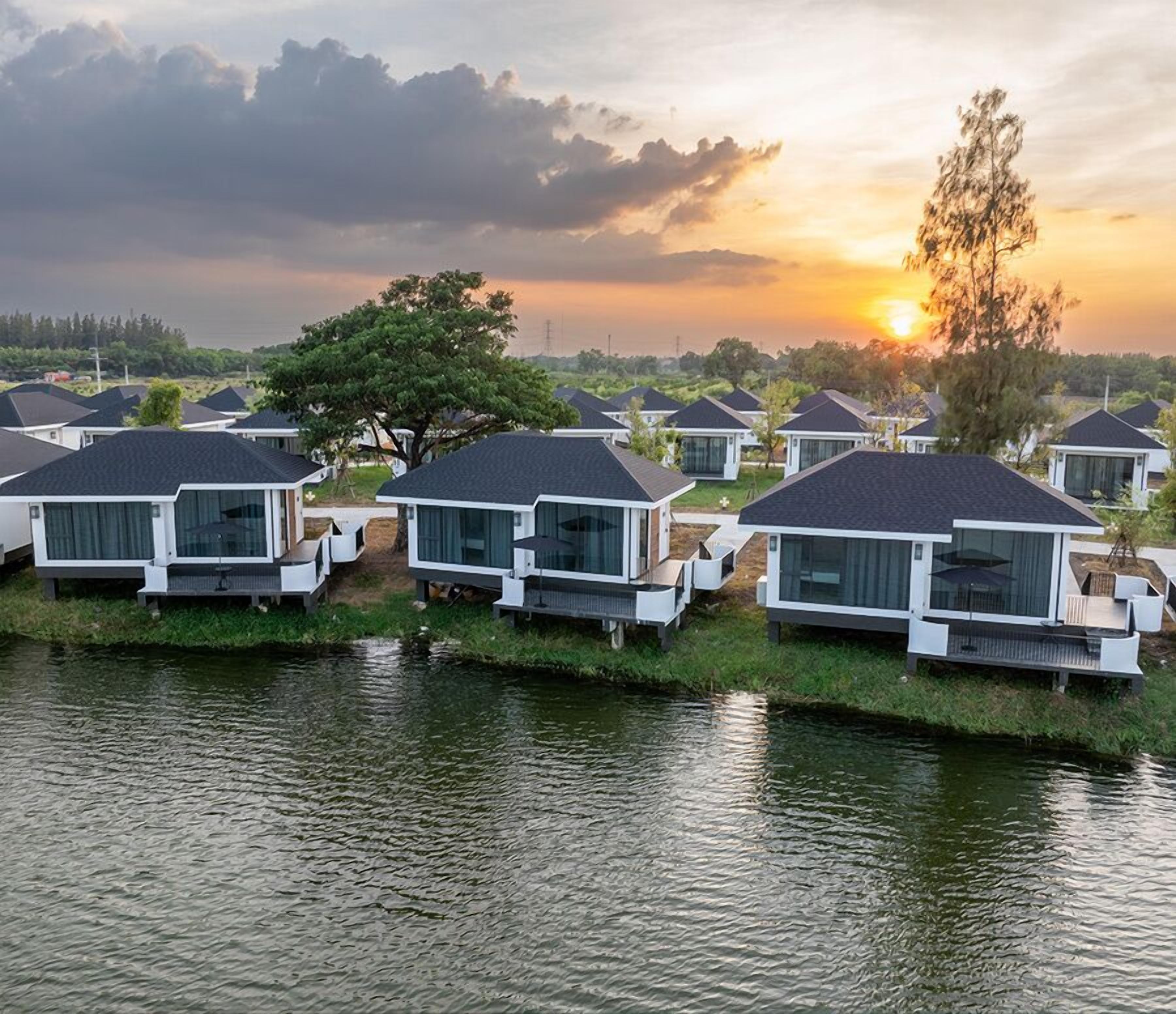 A series of modern, single-story cabins with large windows, situated on stilts over a body of water, with a sunset in the background.