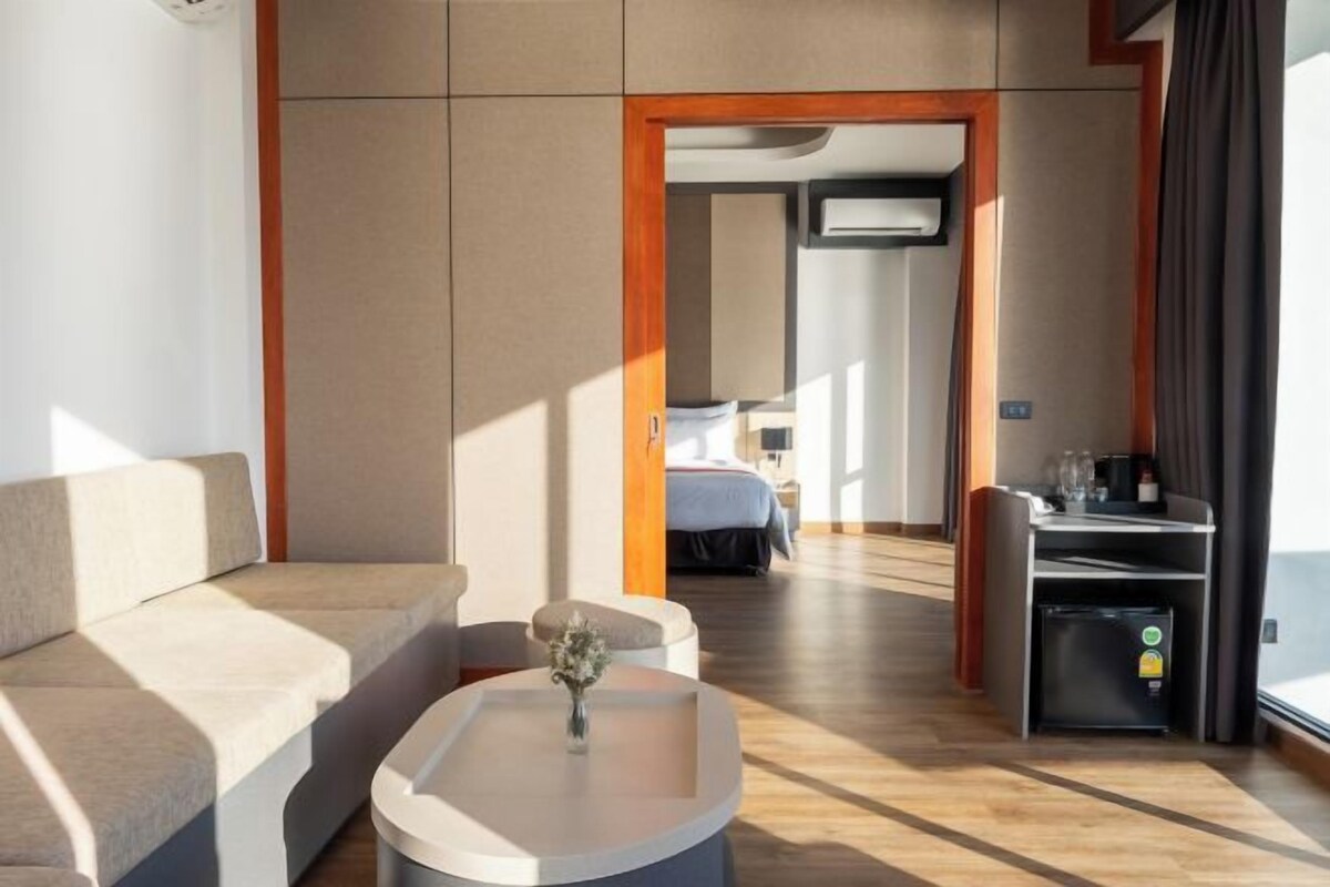 Una habitación de hotel moderna con sofá, una mesita con un florero, una kitchenette y una cama al fondo.