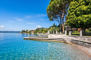 A clear lake with a stone dock, trees, and buildings in the background. - Roby Loft-direct Lake Access/beach (Gardone Riviera)