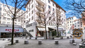A multi-story building with a commercial ground floor, a central open space with bollards, and a clear sky. - Novos Contrada Apartments (Lugano)