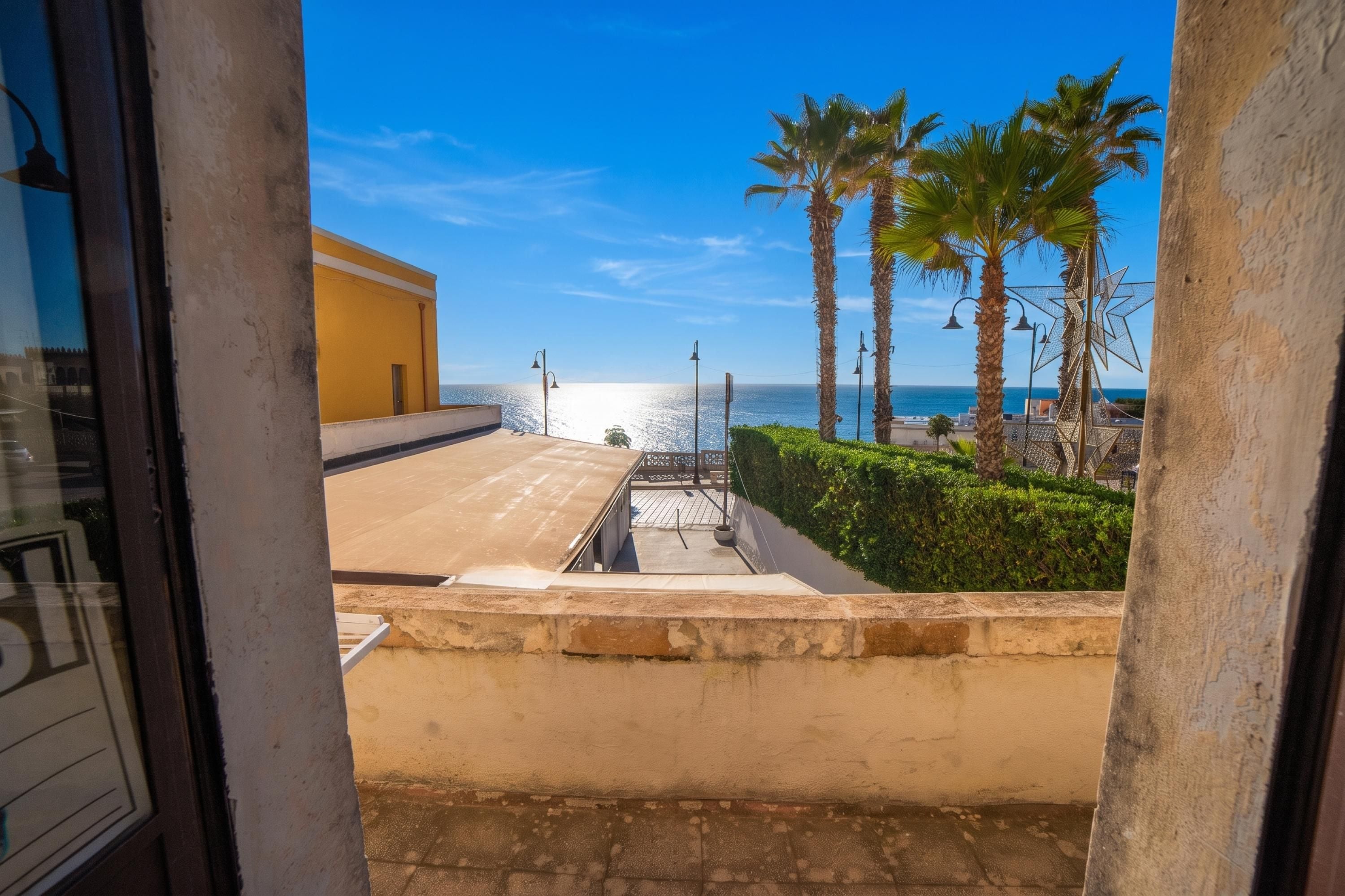 A balcony with a view of the sea, palm trees, and a clear sky.
