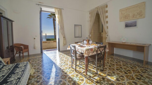 Una stanza con un tavolo da pranzo, sedie, un divano e vista sul mare attraverso una porta aperta.