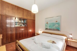 A hotel room with a wooden wardrobe, a bed with white linens, a painting of a ship on the wall, and a bedside table with a lamp. - Casa Virginia (Laveno Mombello)