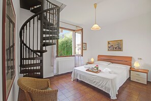 A bedroom with a bed, a nightstand, a bedside lamp, a wooden headboard, a wicker chair, a spiral staircase, and a window with a view of greenery. - Casa Virginia (Laveno Mombello)