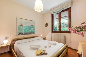 A neatly made bed with white linens, a bedside lamp, a framed picture on the wall, and a window with a view of greenery. - Casa Virginia (Laveno Mombello)