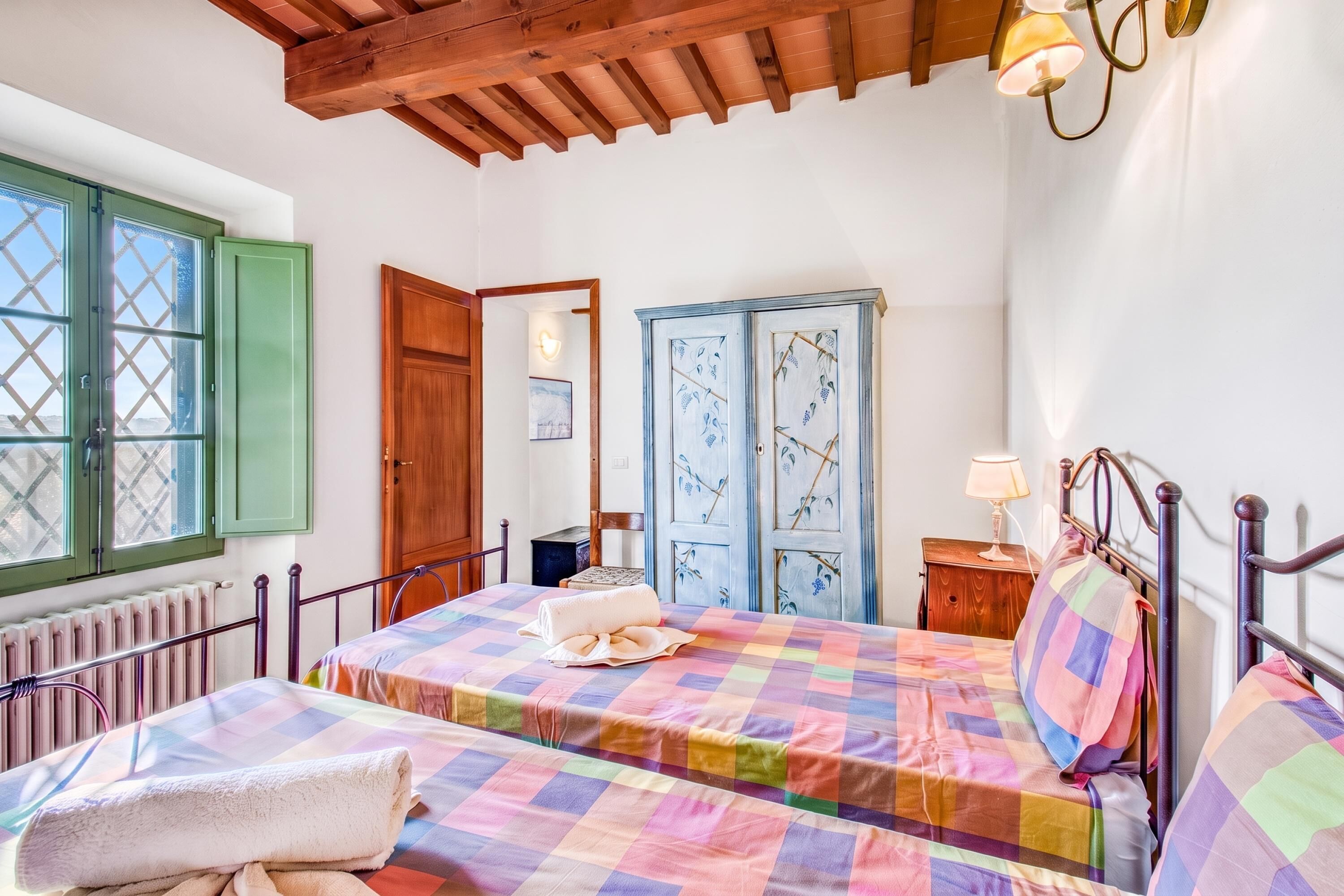 A bedroom with two beds, a wooden ceiling, a radiator, and a window with green shutters.