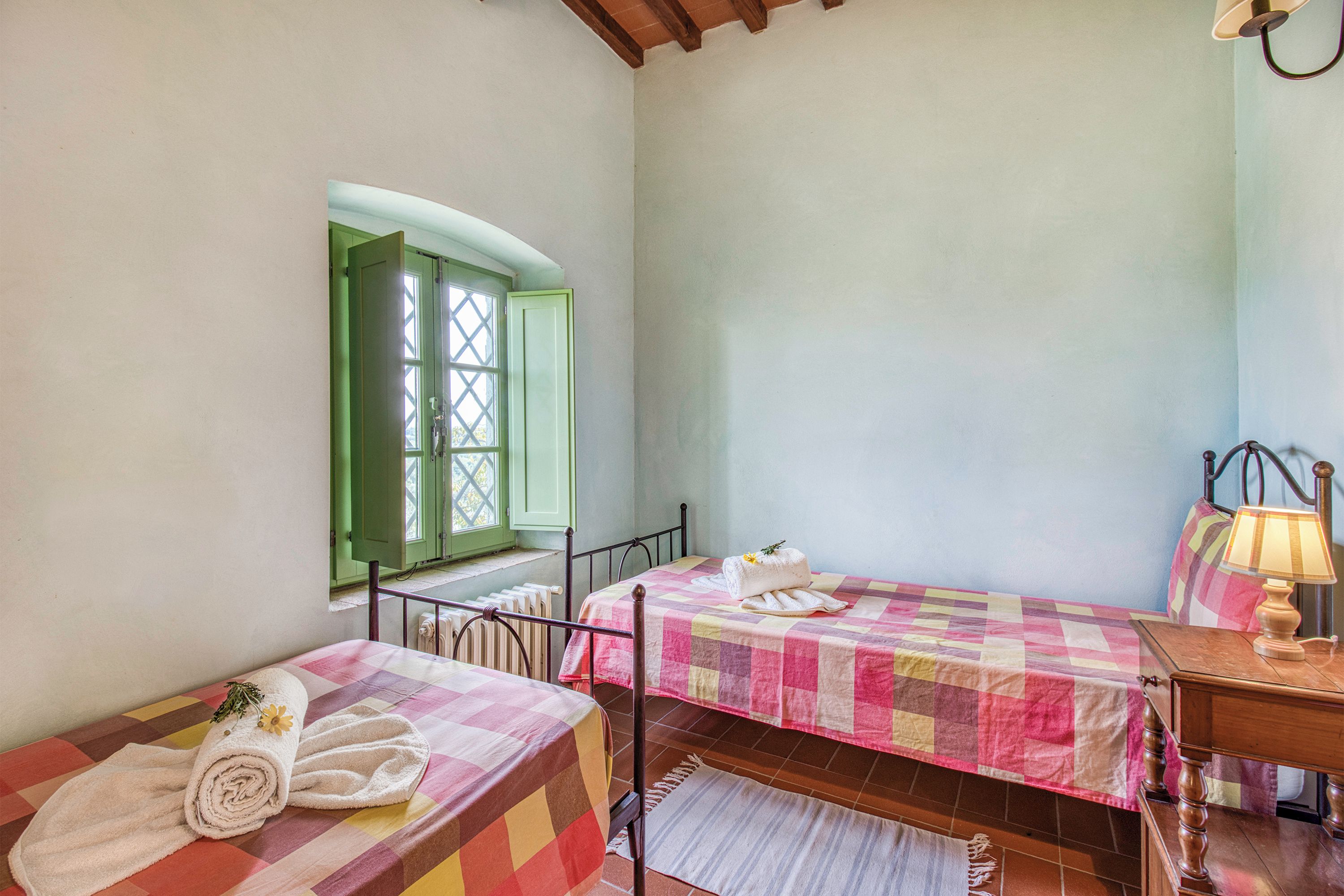 A bedroom with two beds, a window with green shutters, a radiator, and a bedside table with a lamp.