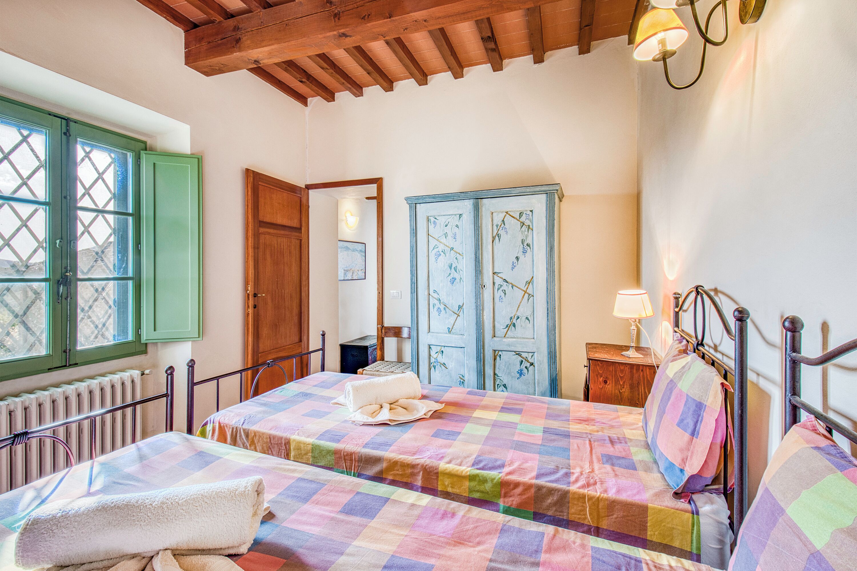 A bedroom with a checkered bedspread, a wooden door, a radiator, and a window with green shutters.