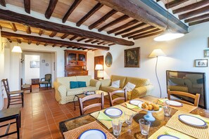 Un coin repas avec une table en bois dressée pour quatre personnes, entourée de chaises. On aperçoit en arrière-plan un canapé avec des coussins, un téléviseur posé sur un meuble et une armoire en bois.