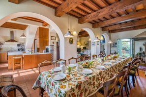 Une vaste salle à manger avec une longue table dressée pour un repas, des chaises en bois et une cuisine en arrière-plan.
