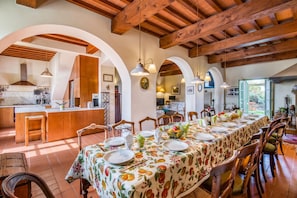 A spacious dining area with a long table set for a meal, wooden beams on the ceiling, and a kitchen in the background.