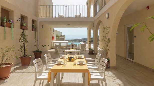 A sunlit outdoor dining area with a table set for six, surrounded by white chairs, potted plants, and a view of the sea. - Ta'Battista B&B with pool Ix-Xwejni (Xewkija)