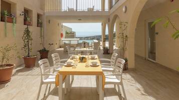 A sunlit outdoor dining area with a table set for six, surrounded by white chairs, potted plants, and a view of the sea.