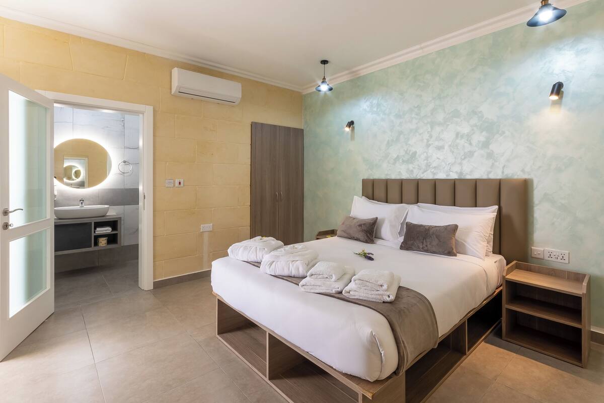 A modern bedroom with a large bed, bedside tables, a bench, and a bathroom with a sink and mirror.