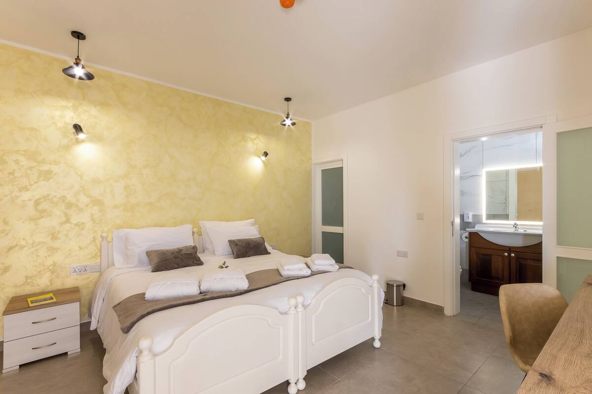 A bedroom with a bed, bedside tables, a mirror, and a bathroom visible through an open door.