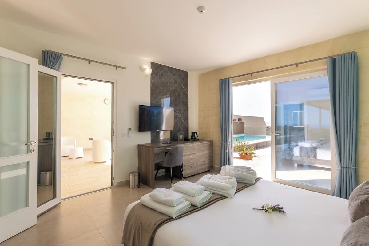 A hotel room with a bed, a desk with a TV, a view of the swimming pool, and a balcony with blue curtains.