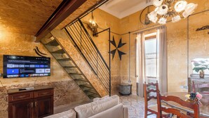 A living room with a staircase, a flat-screen TV mounted on the wall, a wooden cabinet, a sofa, and a dining table with chairs. - Ta'Battista B&B with pool Ix-Xwejni (Xewkija)