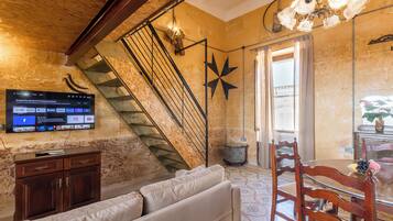 A living room with a staircase, a flat-screen TV mounted on the wall, a wooden cabinet, a sofa, and a dining table with chairs.