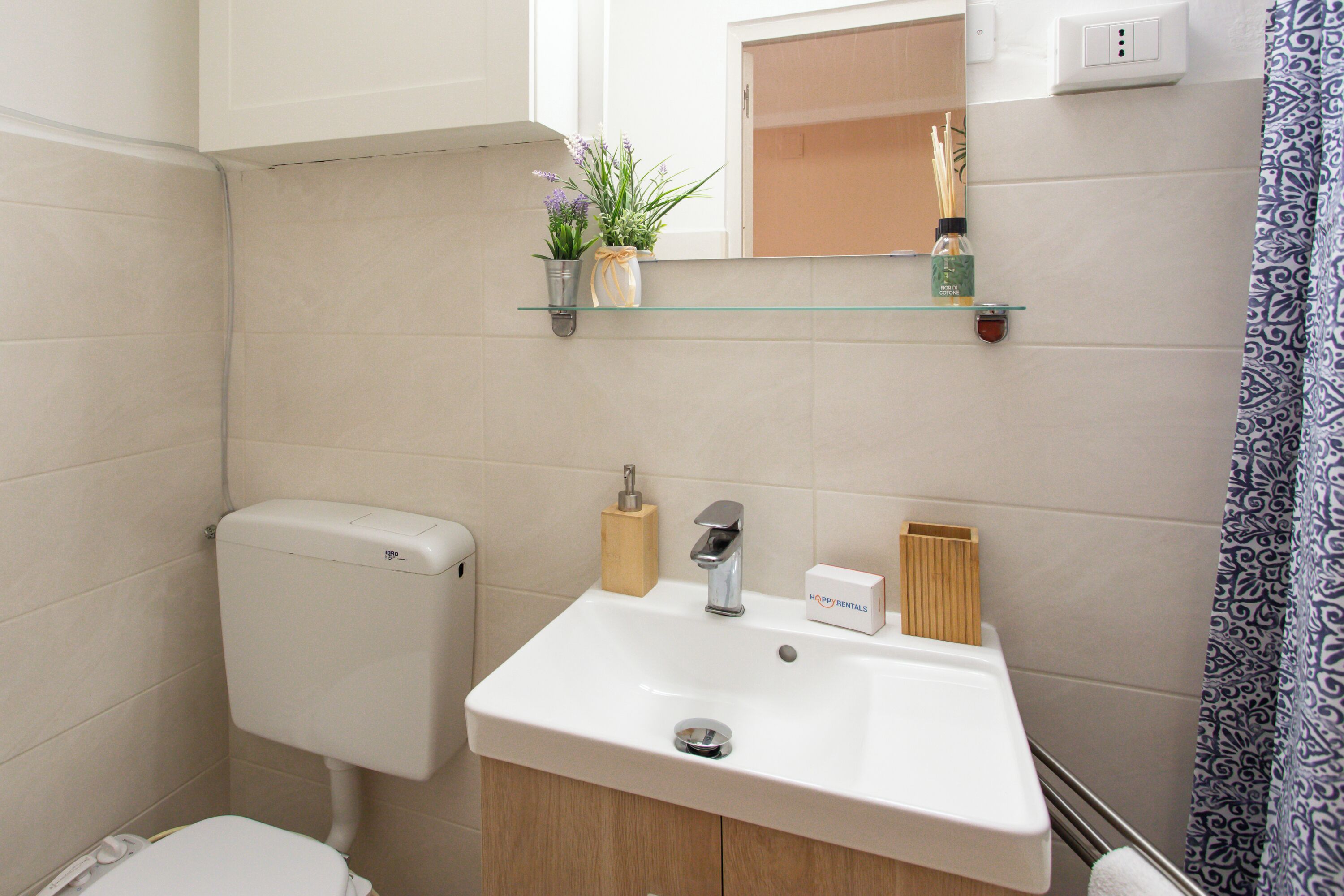 A bathroom with a toilet, sink, and mirror. There are potted plants on the wall and a shower curtain.