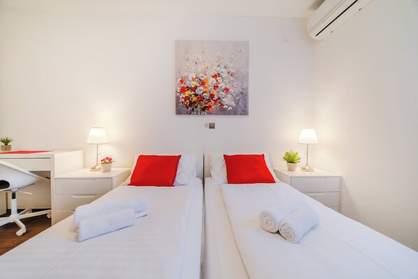 A hotel room with two beds, red pillows, white linens, a desk, a chair, a lamp, a potted plant, and a painting on the wall.