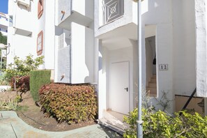 A white building with a door marked "12" and a small garden area with bushes and a hose.