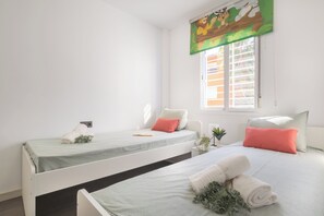 A room with two beds, each with a pillow and a rolled towel. There is a window with blinds and a curtain with a jungle scene.