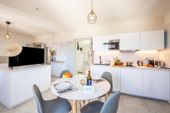 A modern kitchen with white cabinets, a dining table with chairs, a television, and a kitchen island with a fruit bowl and wine bottle. - Tesori Della Vetta (Lugano)