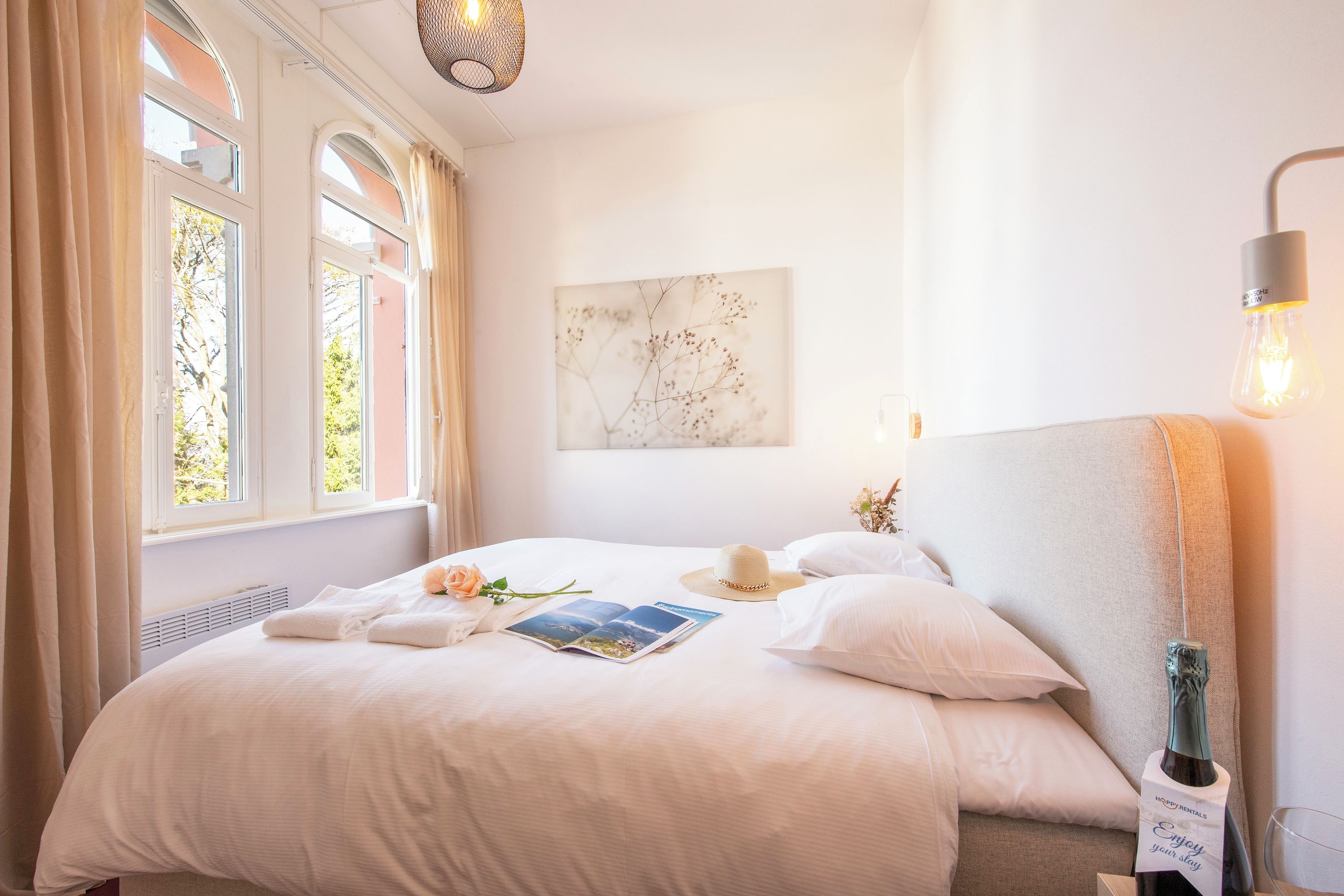 Ein helles Schlafzimmer mit einem großen Bett, einem Nachttisch mit einer Champagnerflasche und einer Wandleuchte.