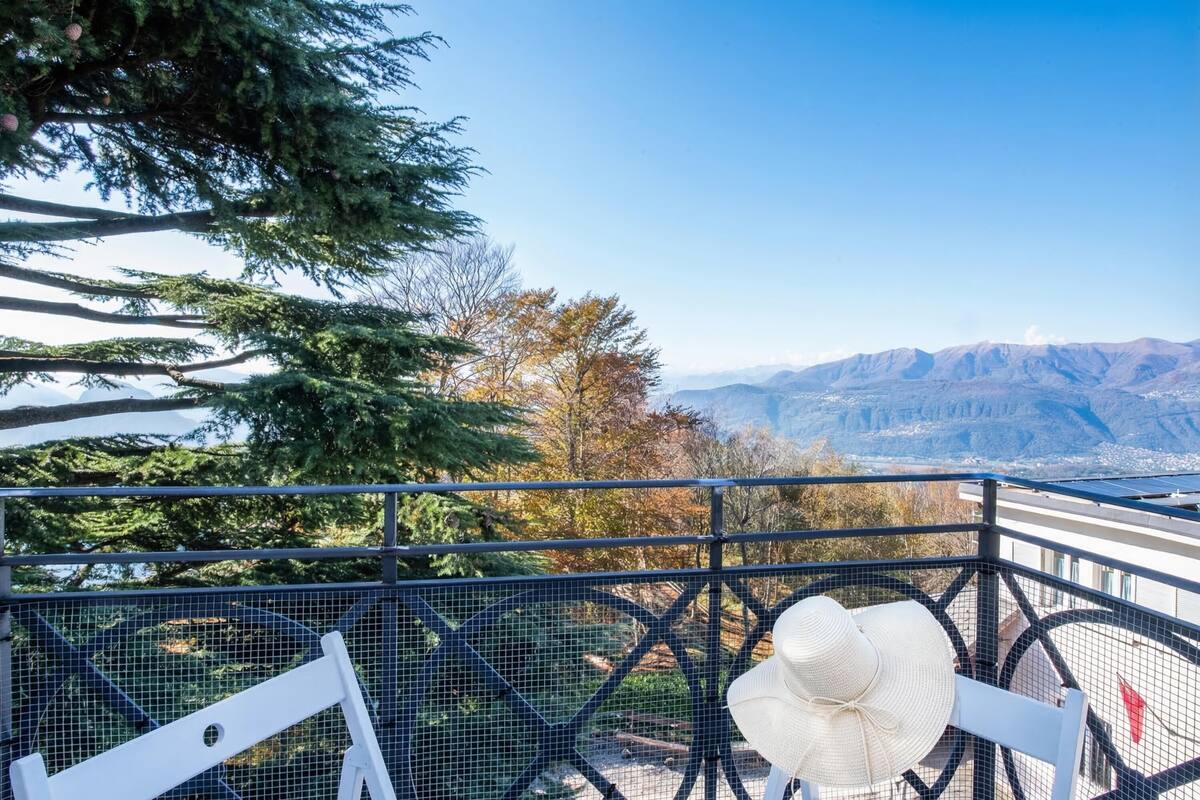 Un balcon avec une chaise blanche, un chapeau blanc et une vue sur les montagnes et les arbres.