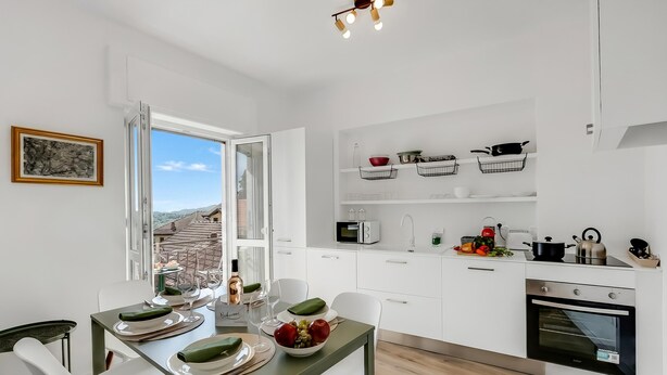 A modern kitchen with white cabinets, a dining table set with plates and glasses, and a view through an open window. - Luce Lake View on Stresa Hill (Stresa)