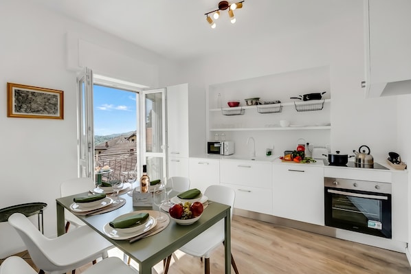 A modern kitchen with white cabinets, a dining table set with plates and glasses, and a view through an open window. - Luce Lake View on Stresa Hill (Stresa)