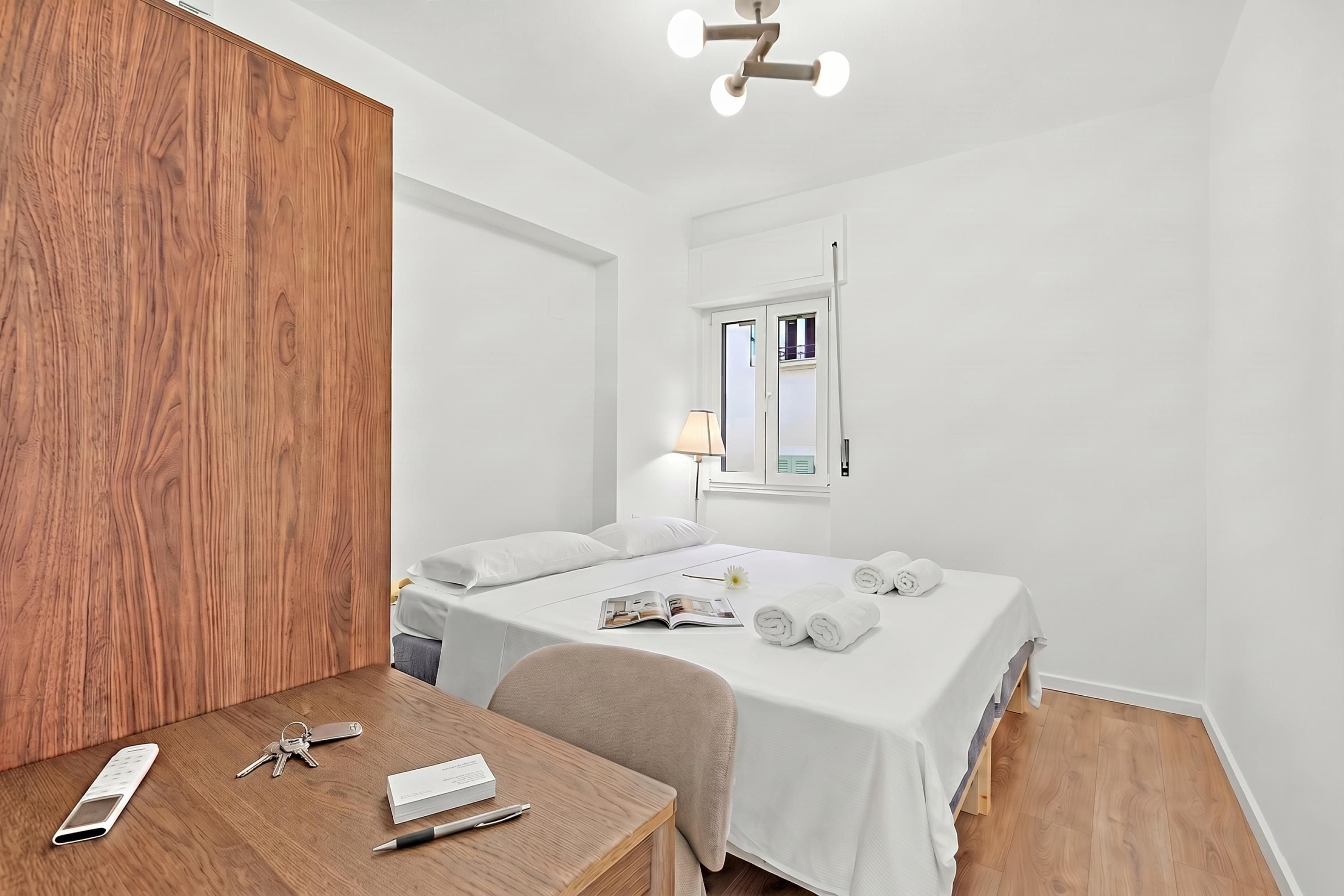 A hotel room with a wooden accent wall, a bed with white linens, a desk with a phone and keys, and a lamp.