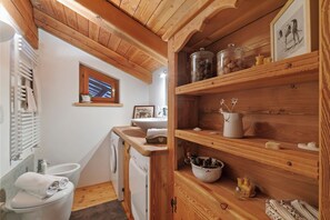 A compact bathroom with a toilet, sink, and a wooden shelf with jars and a picture frame.