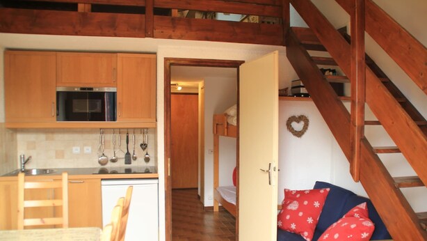 A compact kitchen with wooden cabinets, a microwave, and a sink. A dining table and chairs are visible. A staircase leads to a loft area with a bed and a heart-shaped decoration on the wall.