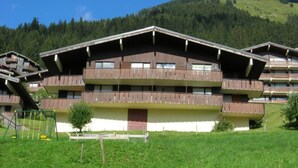 A multi-story chalet with balconies, set in a mountainous area with a grassy foreground and a forested hill in the background.