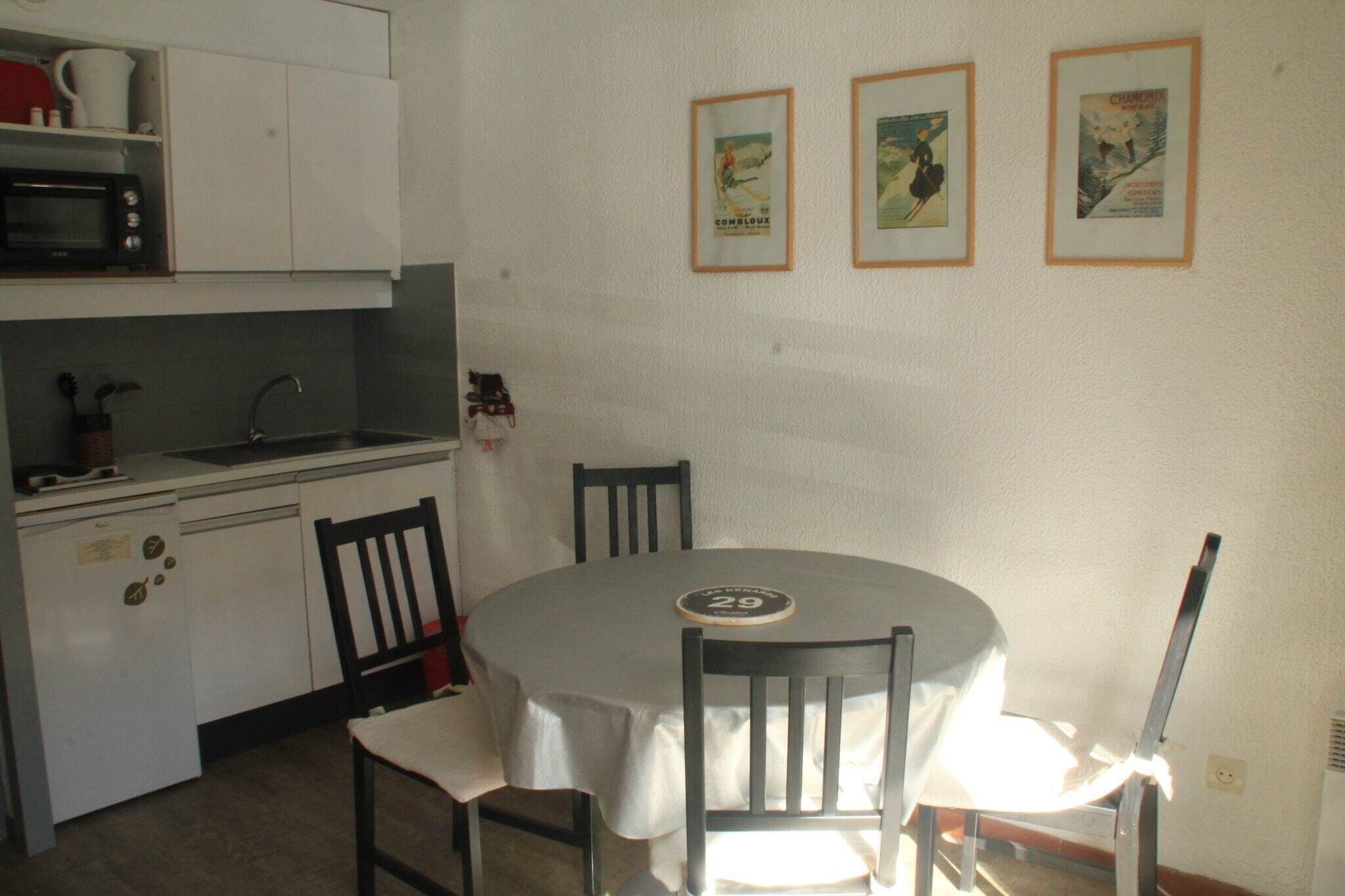 A compact kitchen with a microwave, sink, and cabinets. There are two framed pictures on the wall and a round table with chairs.