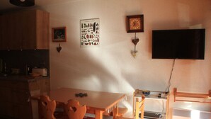 A kitchen with wooden cabinets, a dining table with chairs, a framed picture, a clock, and a TV mounted on the wall. - Apartment in Chatel in ski resort (Châtel)