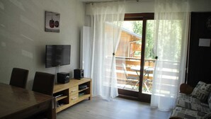 A living room with a wooden dining table, chairs, a TV on a stand, a sofa, and a sliding glass door leading to an outdoor area with a wooden deck. - Apartment in Portes du Soleil (Châtel)