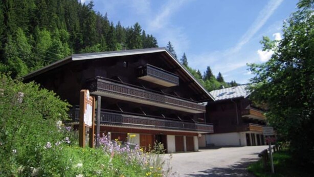 Ein Chalet mit geneigtem Dach und hölzernen Balkonen, umgeben von üppiger Vegetation und einem bewaldeten Hintergrund.