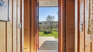 A wooden interior leading to a wooden deck with a view of a garden and a building in the distance. - Cozy cottage with a beautiful view. (Tranekær)
