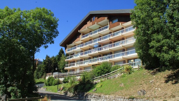Un hotel de varios pisos con balcones, ubicado en una colina con árboles y cielo despejado.