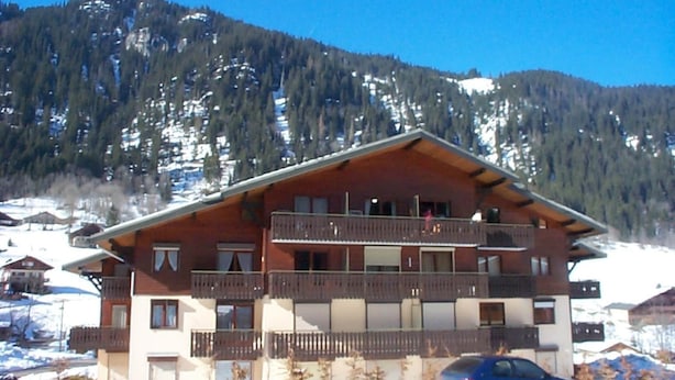 A multi-story building with balconies, a car parked in front, and a mountainous backdrop.