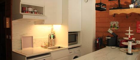 A compact kitchen with white cabinetry, a microwave, a built-in washing machine, and a wooden wall with a clock and a shelf.