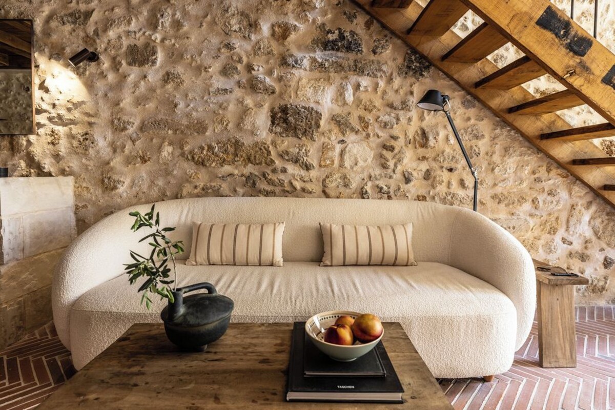 Une pièce chaleureuse avec un mur en pierre, un canapé incurvé, une petite table sur laquelle se trouve un saladier rempli de fruits et une lampe.